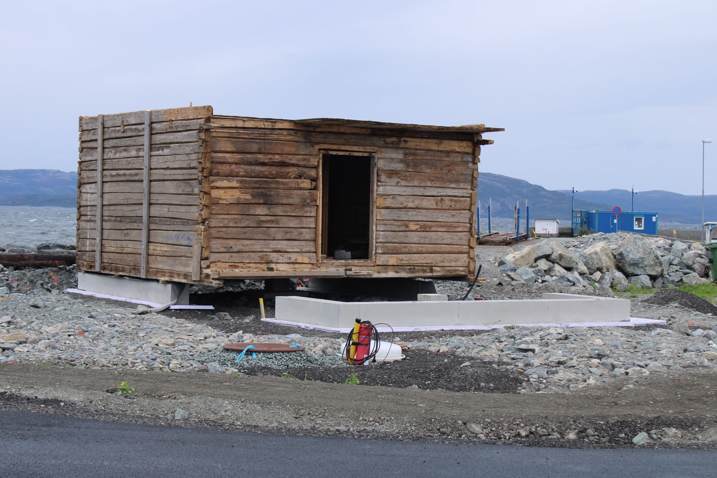Stabburet gjenreist ved sjøen på en ny betongsåle, med steinfylling, brakker og fjell i bakgrunnen.