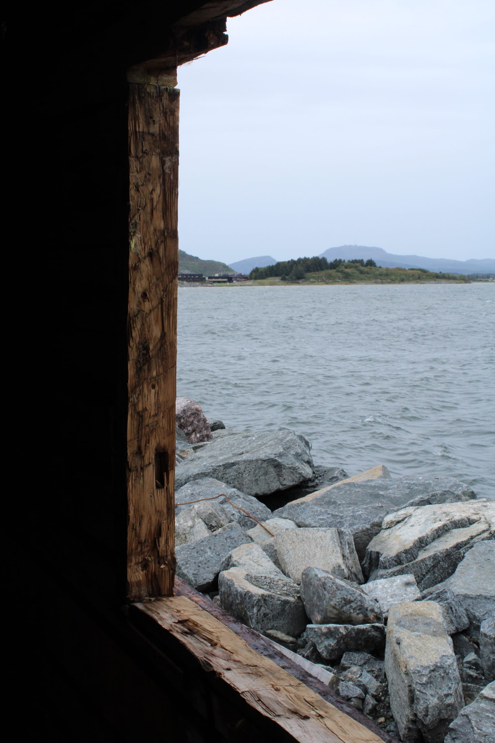 Utsikt ut gjennom den mørke åpningen i stabburet: en tømmerstolpe i kanten, steinfyllingen utenfor og sjøen med landet bak.
