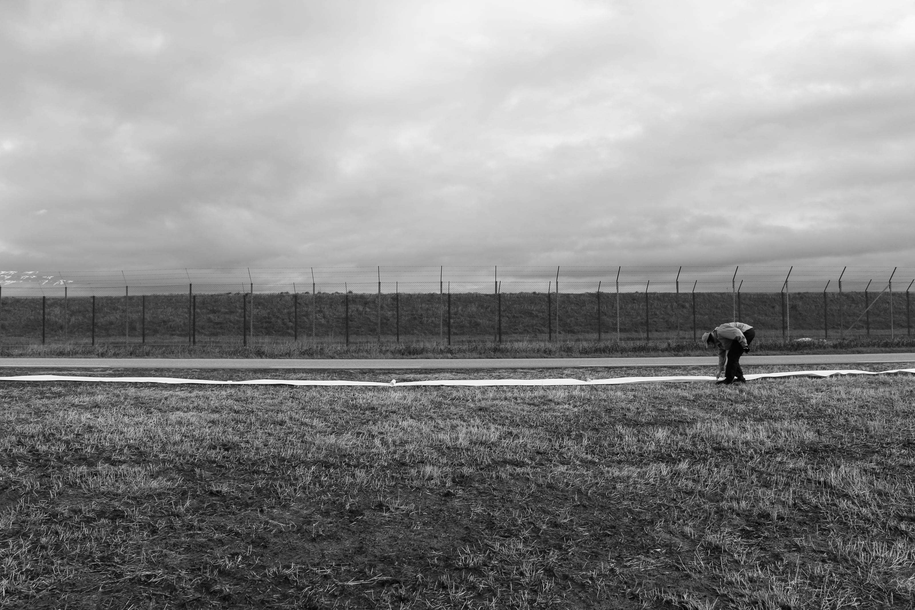 En person bøyer seg ned og legger en lang, hvit remse langs kanten av rullebanen, med flyplassgjerdet og en lav horisont bak.