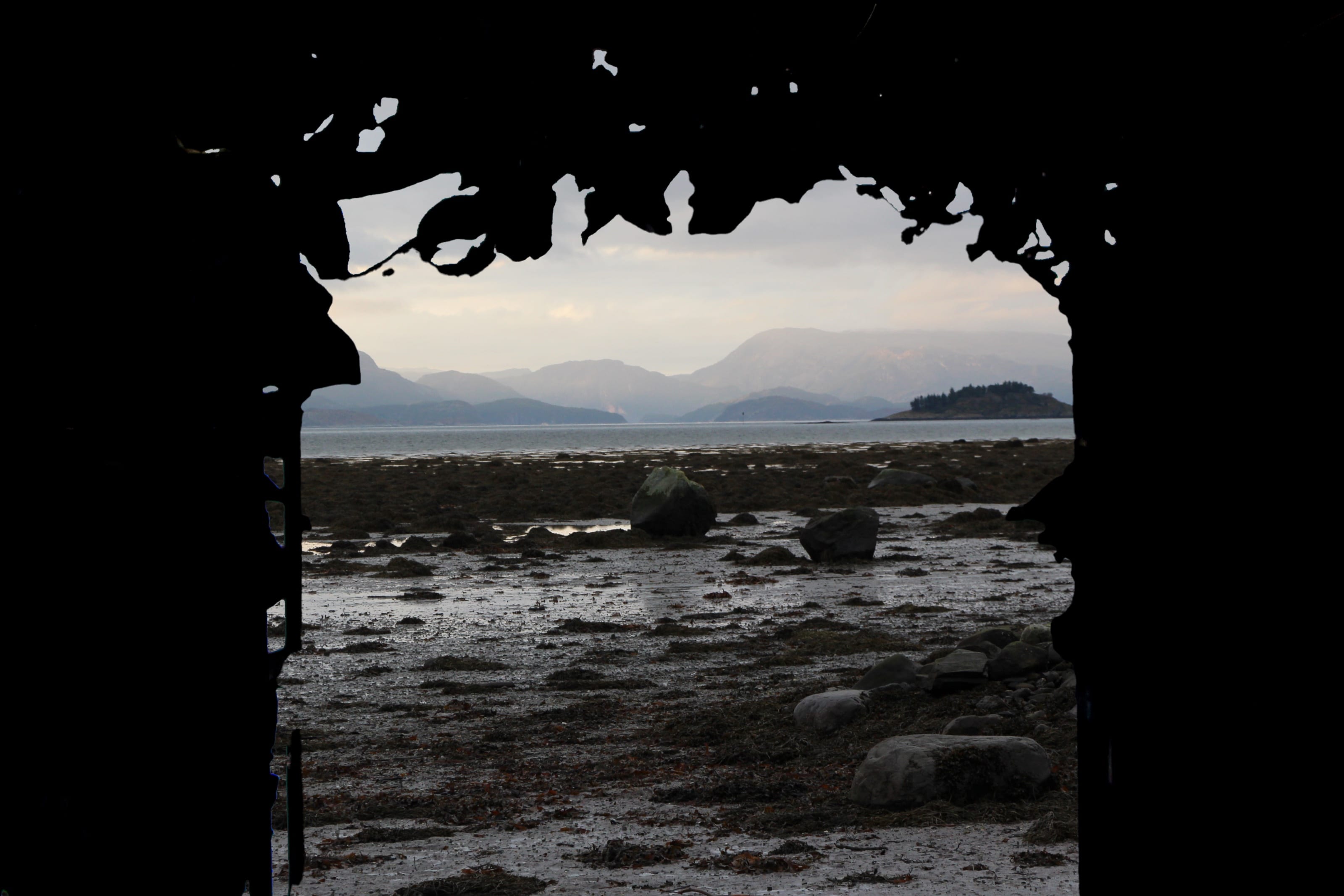 Fotografi tatt innenfra Tangportalen: den mørke tangkledde åpningen rammer inn fjæra med steiner og tang, fjorden og fjellene bak.