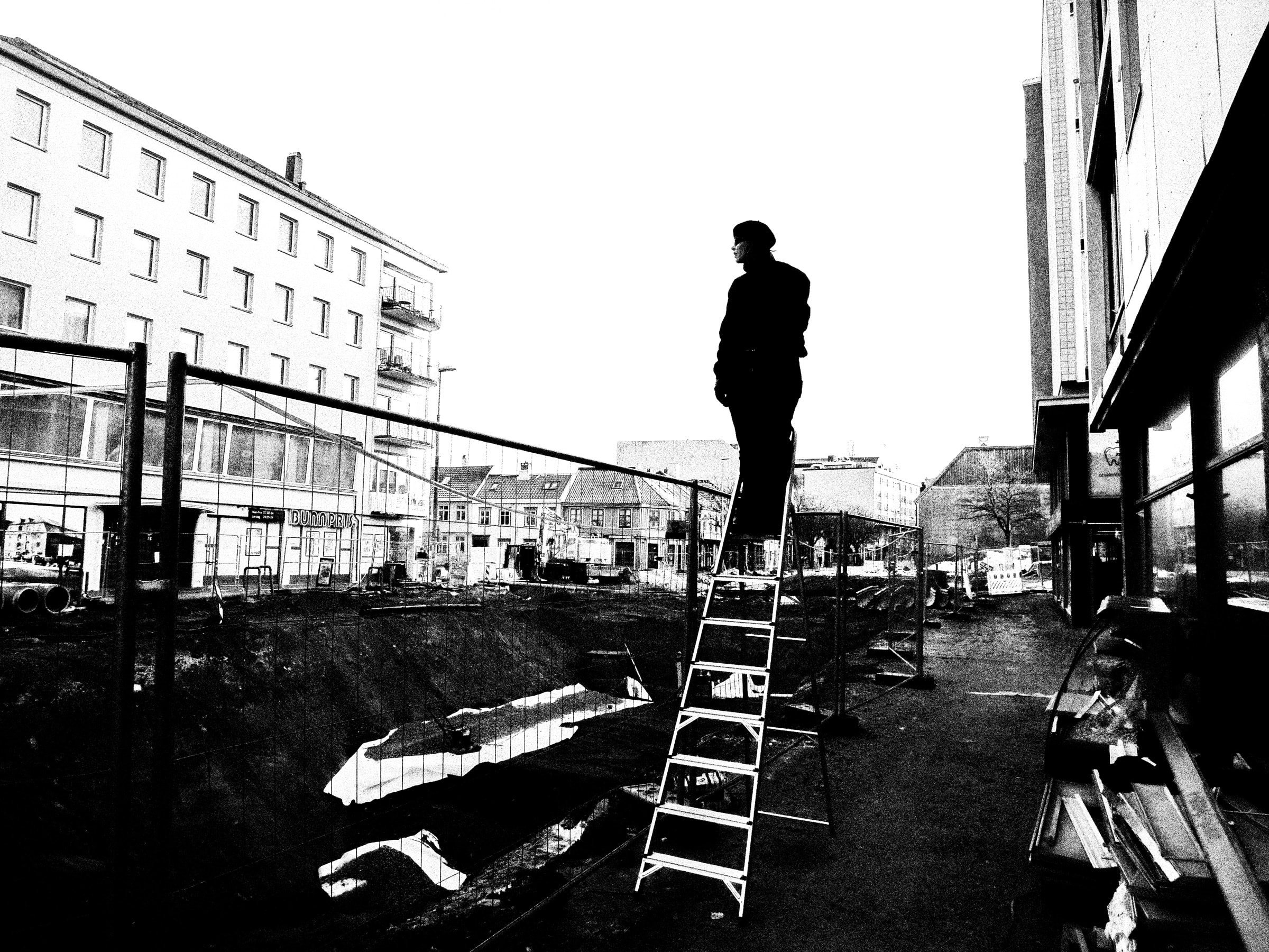 En skikkelse i sort står oppe på en gardintrapp på en byggeplass, i silhuett mot en hvit himmel, med gjerder og gravemasser rundt seg.