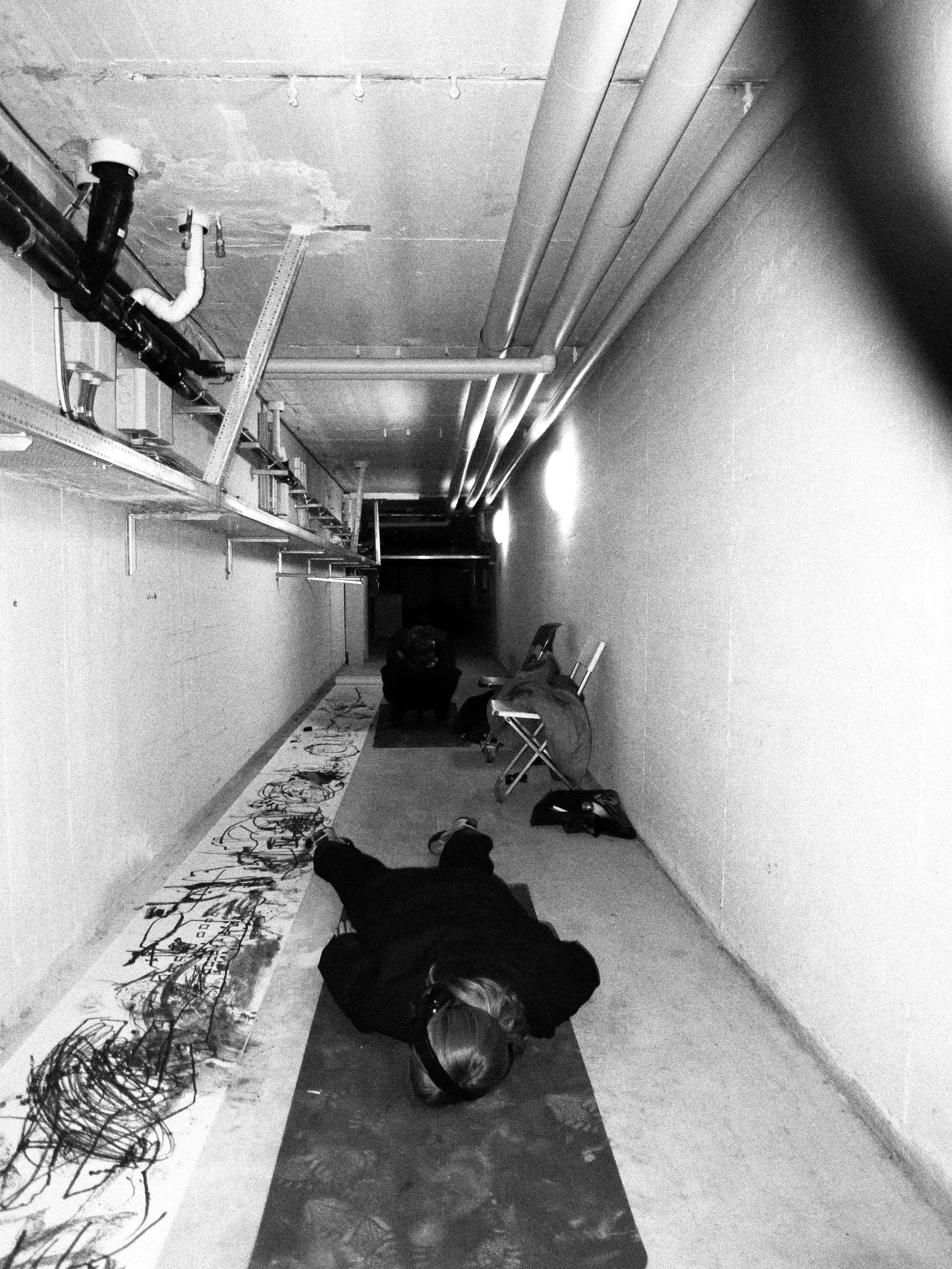 Kjellergangen ovenfra: den ene skikkelsen ligger utstrakt på gulvet, den andre sitter sammenkrøpet lenger inne, og den lange tegningen ligger utrullet langs veggen.