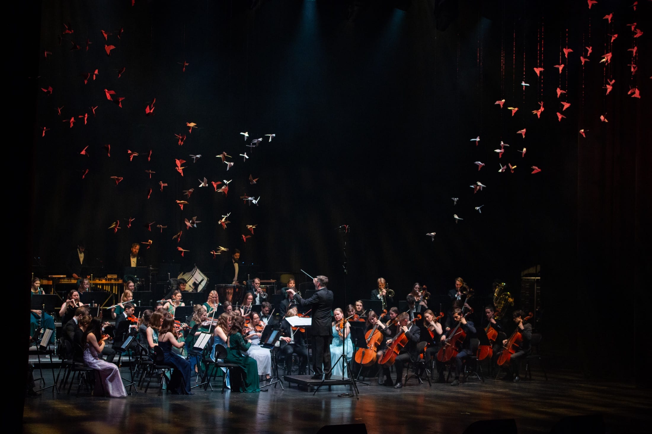 Et symfoniorkester som spiller på scenen under den hengende flokken av papirfugler.